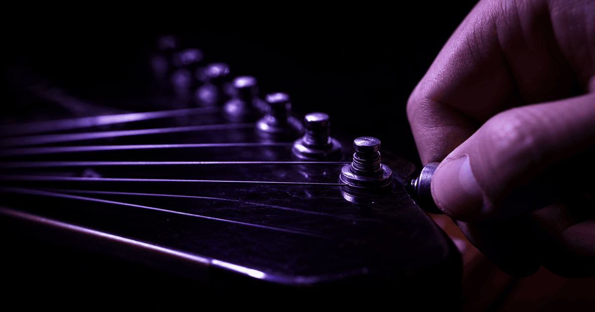 A hand turning a tuning peg on a guitar headstock in a dark studio, the strings receding into shadow under a raking purple light